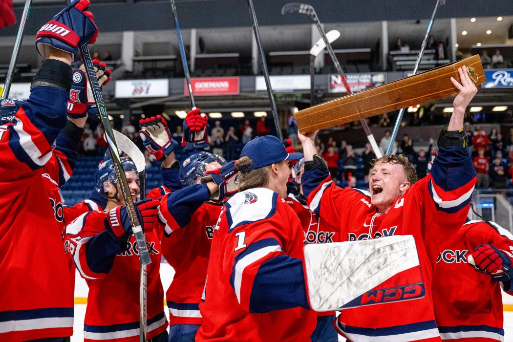 A hockey player hoists a trophy above his head while surrounded by his teammates.