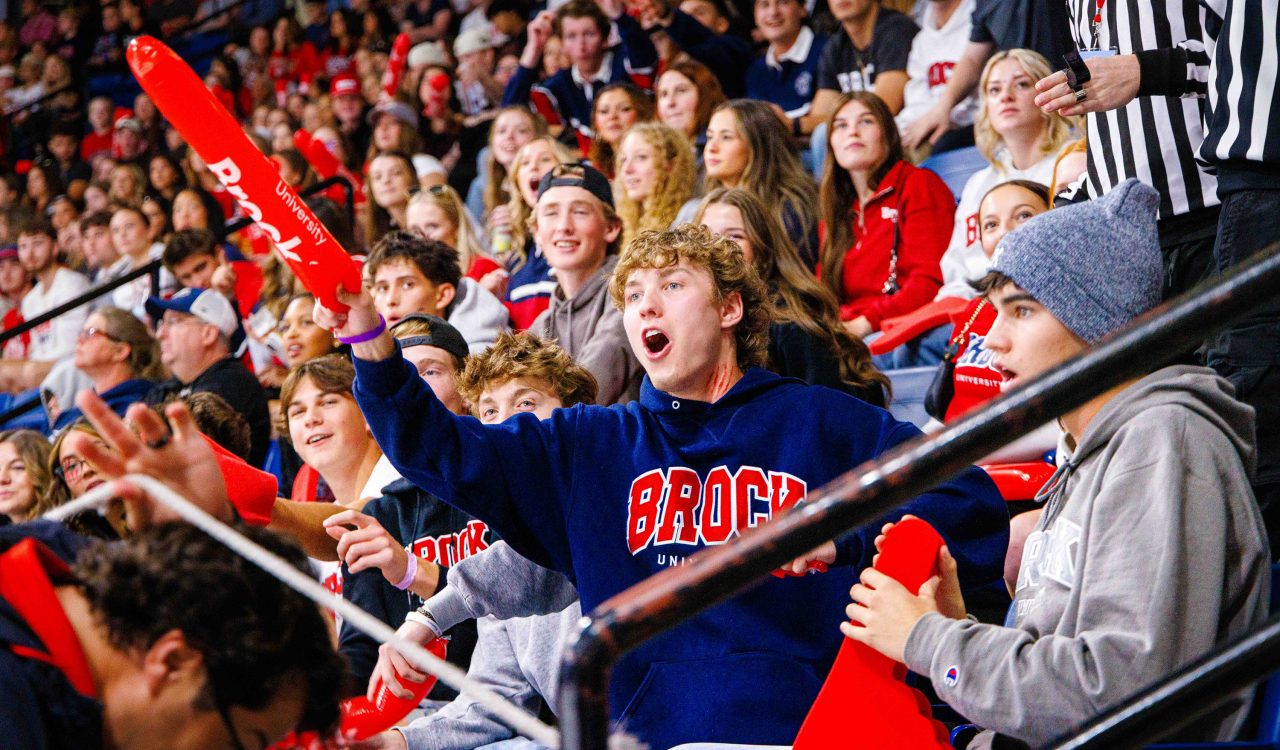 A crowd of people in Brock University gear cheer while sitting in stadium seats.