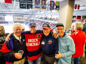 Three older man poses with Boomer the Badger, Brock University's mascot, at a hockey game.
