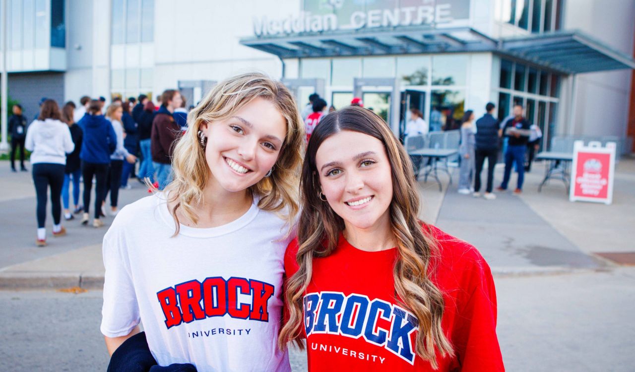 Two women in Brock University t-shirts stand outside the Meridian Centre arena with a crowd in the background.