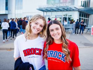 Two women in Brock University t-shirts stand outside the Meridian Centre arena with a crowd in the background.