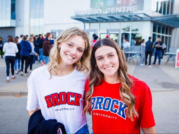 Two women in Brock University t-shirts stand outside the Meridian Centre arena with a crowd in the background.