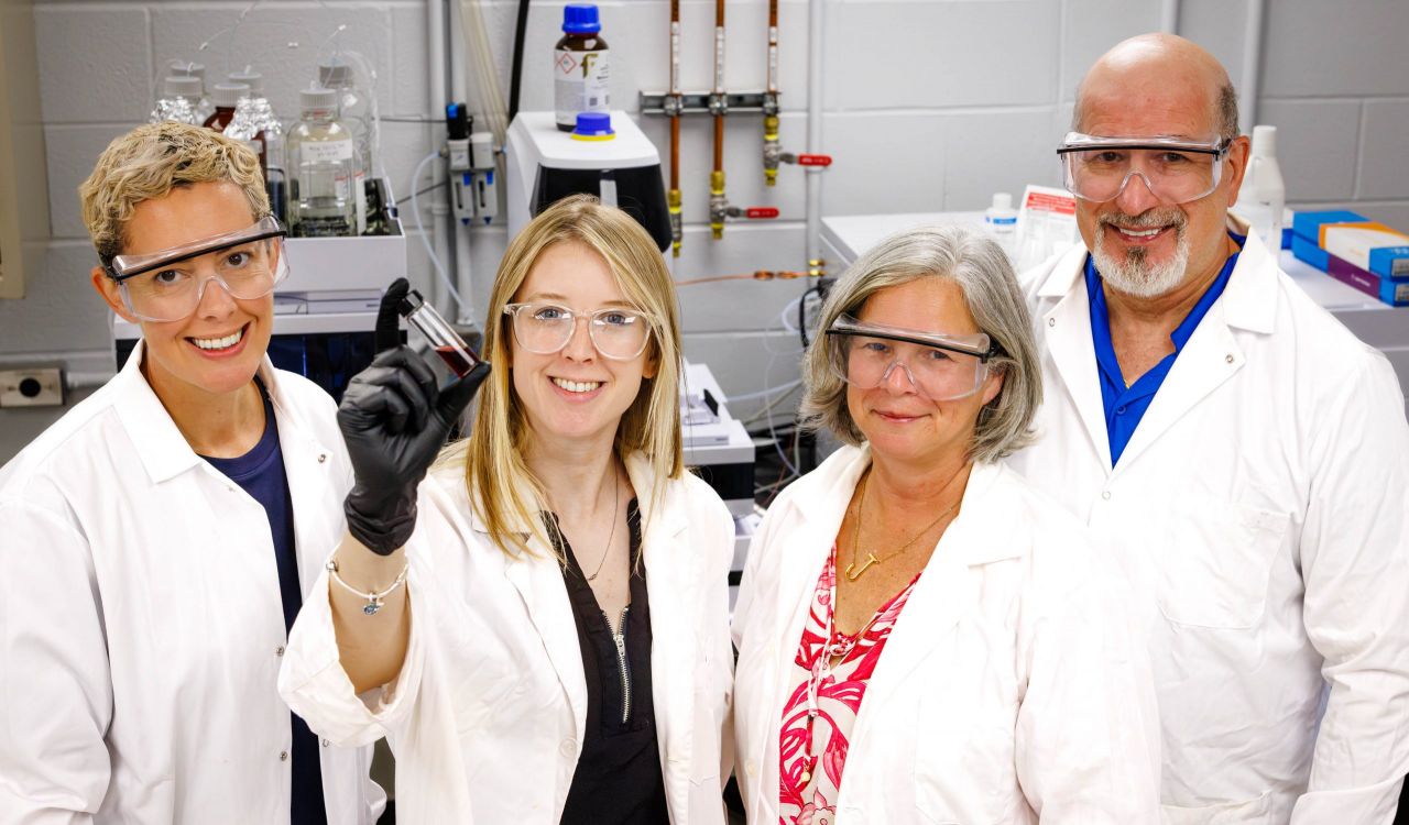 Three women and a man wearing goggles and white lab coats stand in front of mchines in a lab, with one of the women holding up a vial of red liquid.