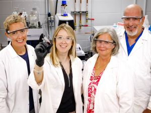 Three women and a man wearing goggles and white lab coats stand in front of mchines in a lab, with one of the women holding up a vial of red liquid.