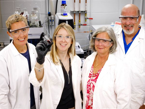 Three women and a man wearing goggles and white lab coats stand in front of mchines in a lab, with one of the women holding up a vial of red liquid.
