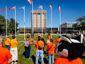 A group of people wearing orange shirts stand outdoors on the Brock campus, facing seven flagpoles and the Schmon Tower.