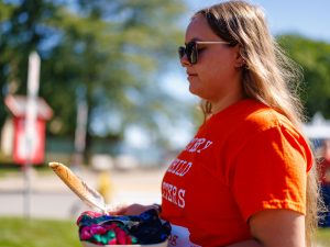 A woman stands outdoors, holding a ribbon in one hand and a bowl in the other. She is wearing sunglasses and an orange Every Child Matters ft-shirt