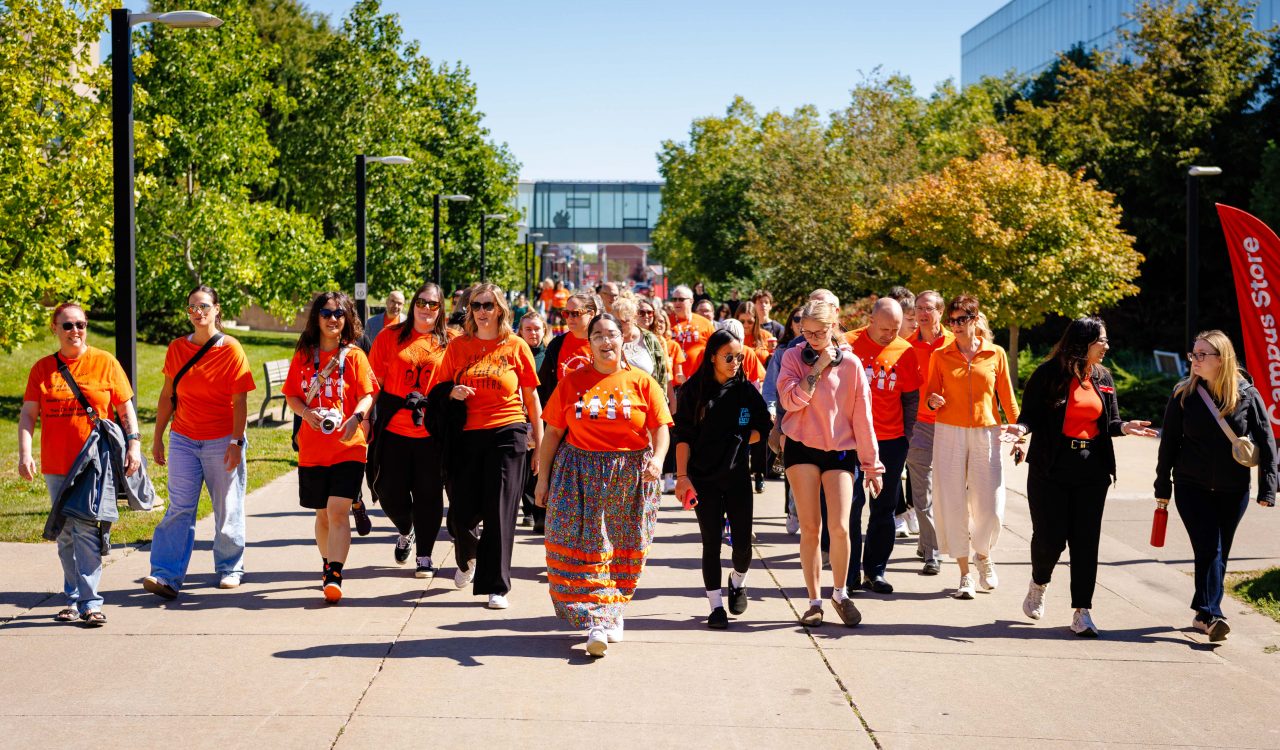 A group of Brock University students, faculty and staff walk together on an outdoor paved pathway with campus buildings visible behind them. Most of the people are wearing orange shirts.