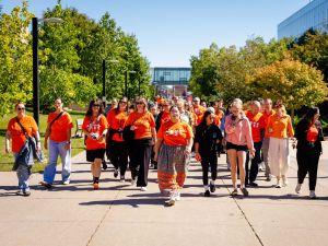 A group of Brock University students, faculty and staff walk together on an outdoor paved pathway with campus buildings visible behind them. Most of the people are wearing orange shirts.