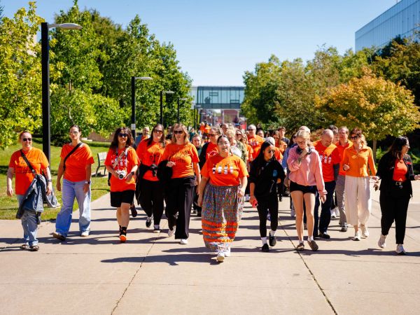 A group of Brock University students, faculty and staff walk together on an outdoor paved pathway with campus buildings visible behind them. Most of the people are wearing orange shirts.