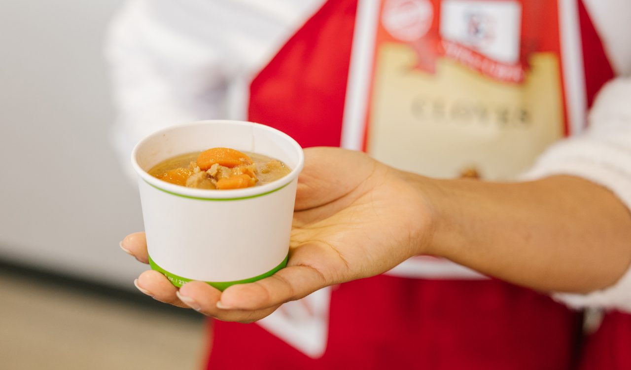 A hand is extended holding a small container of soup.
