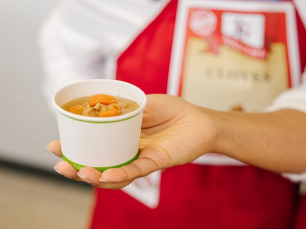 A hand is extended holding a small container of soup.