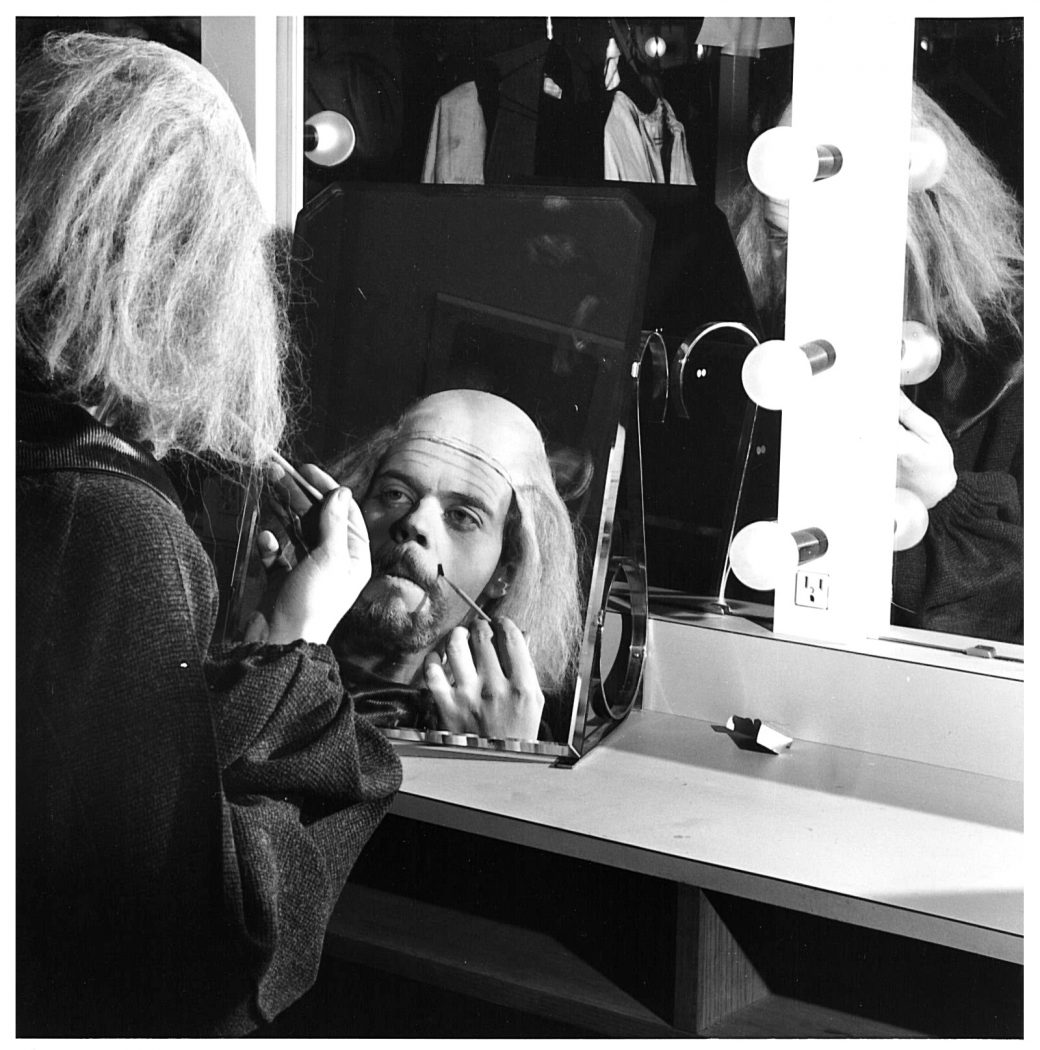 A man puts on stage makeup while looking in a mirror. 