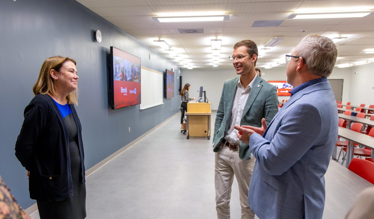 Alexandra Lawson, Sam Oosterhoff and Scott Johnstone stand in a modern classroom, talking beside rows of tables and chairs. Lawson stands on the left facing Oosterhoff and Johnstone, who are engaged in conversation near the front of the room. Large display screens are mounted on the wall behind them.