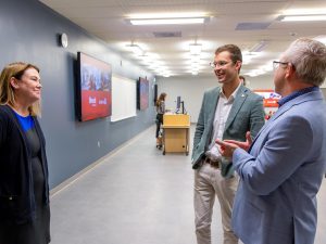 Alexandra Lawson, Sam Oosterhoff and Scott Johnstone stand in a modern classroom, talking beside rows of tables and chairs. Lawson stands on the left facing Oosterhoff and Johnstone, who are engaged in conversation near the front of the room. Large display screens are mounted on the wall behind them.