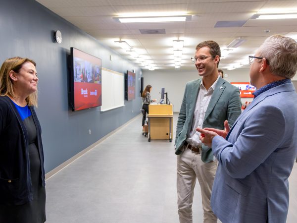Alexandra Lawson, Sam Oosterhoff and Scott Johnstone stand in a modern classroom, talking beside rows of tables and chairs. Lawson stands on the left facing Oosterhoff and Johnstone, who are engaged in conversation near the front of the room. Large display screens are mounted on the wall behind them.