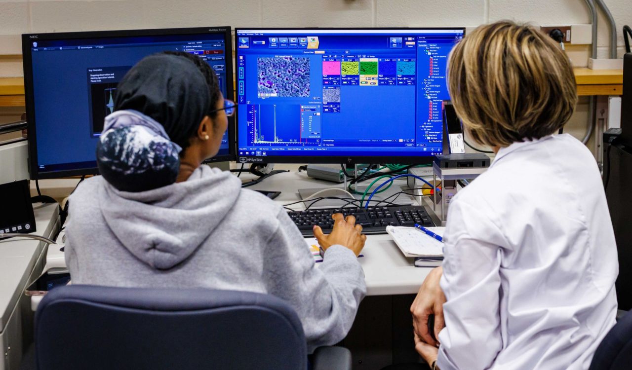 Two women scientists, their backs to the camera, look at a screen displaying graphs and illustrations.
