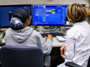 Two women scientists, their backs to the camera, look at a screen displaying graphs and illustrations.