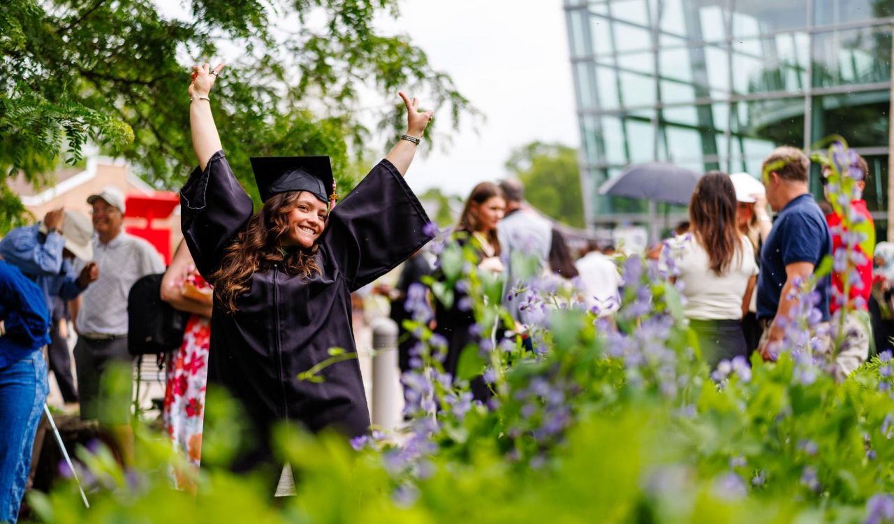 A woman in a graduation robe and cap stands outside with both hands raised above her head.