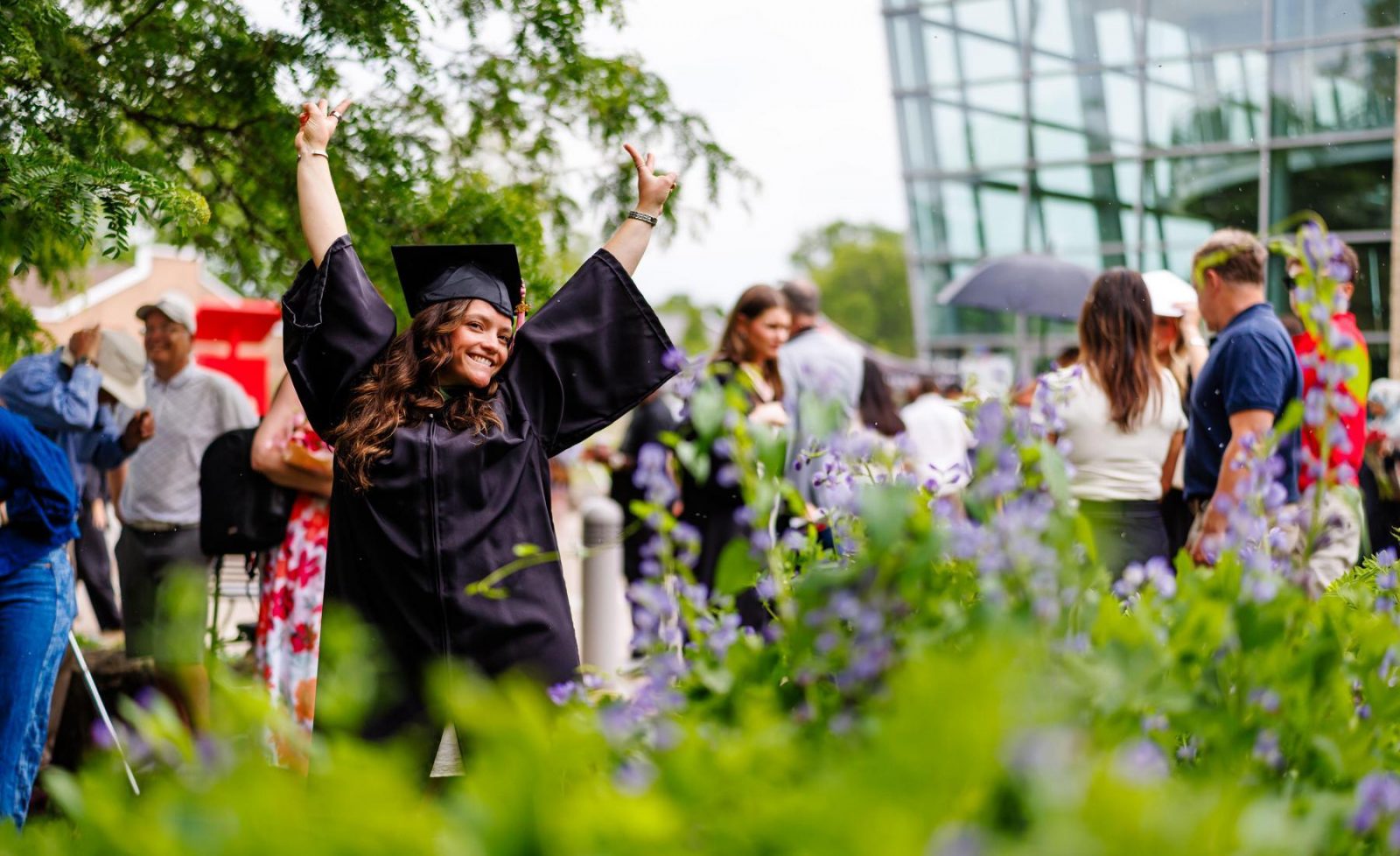A woman in a graduation robe and cap stands outside with both hands raised above her head.