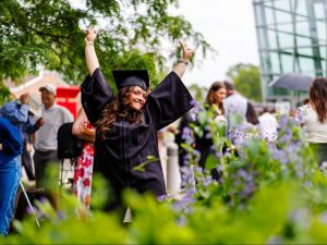A woman in a graduation robe and cap stands outside with both hands raised above her head.