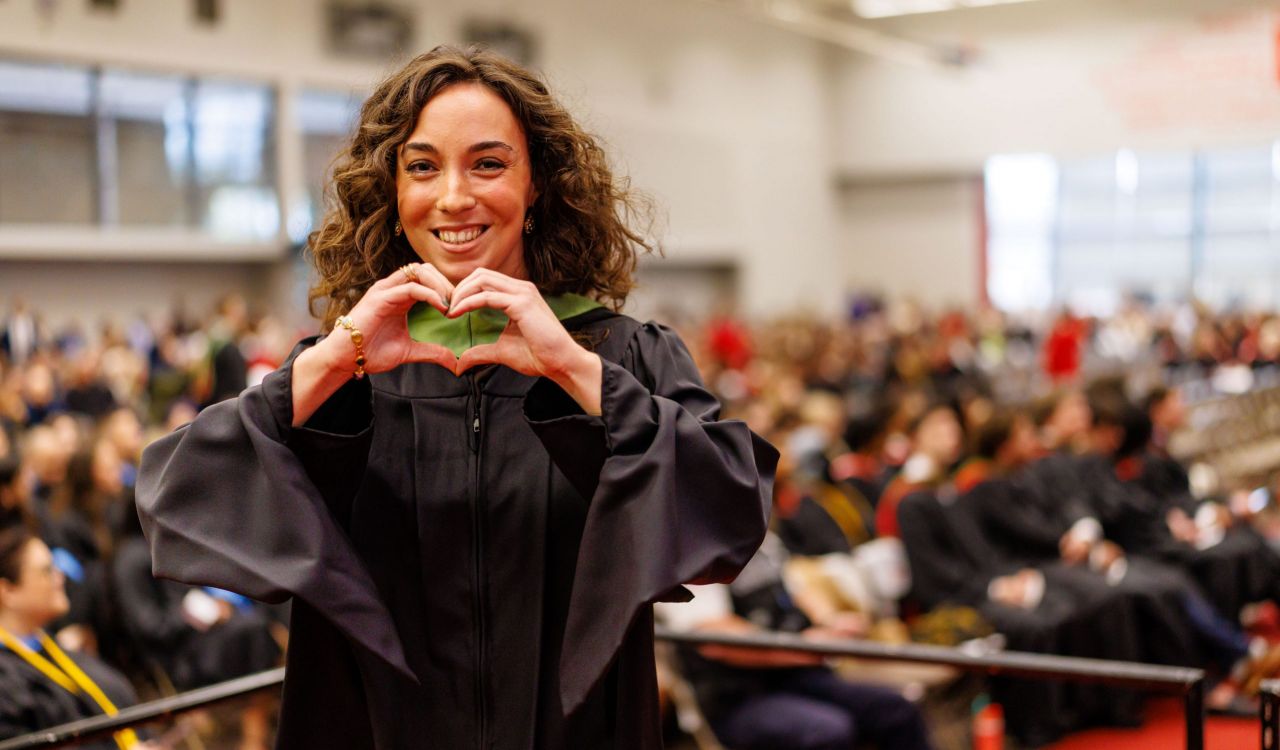 Alyssa Scalera smiles, wearing a black graduation robe with a green hood on stage at Fall Convocation held inside the Ian D. Beddis Gymnasium at Brock University.