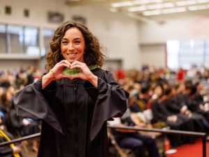 Alyssa Scalera smiles, wearing a black graduation robe with a green hood on stage at Fall Convocation held inside the Ian D. Beddis Gymnasium at Brock University.