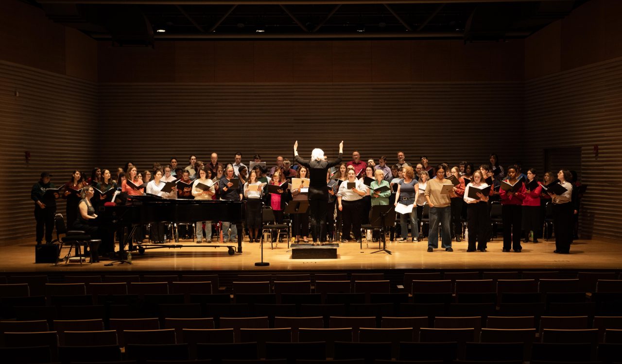 A conductor leads a large group of people who are singing on stage in a professional theatre with auditorium seating in the foreground.
