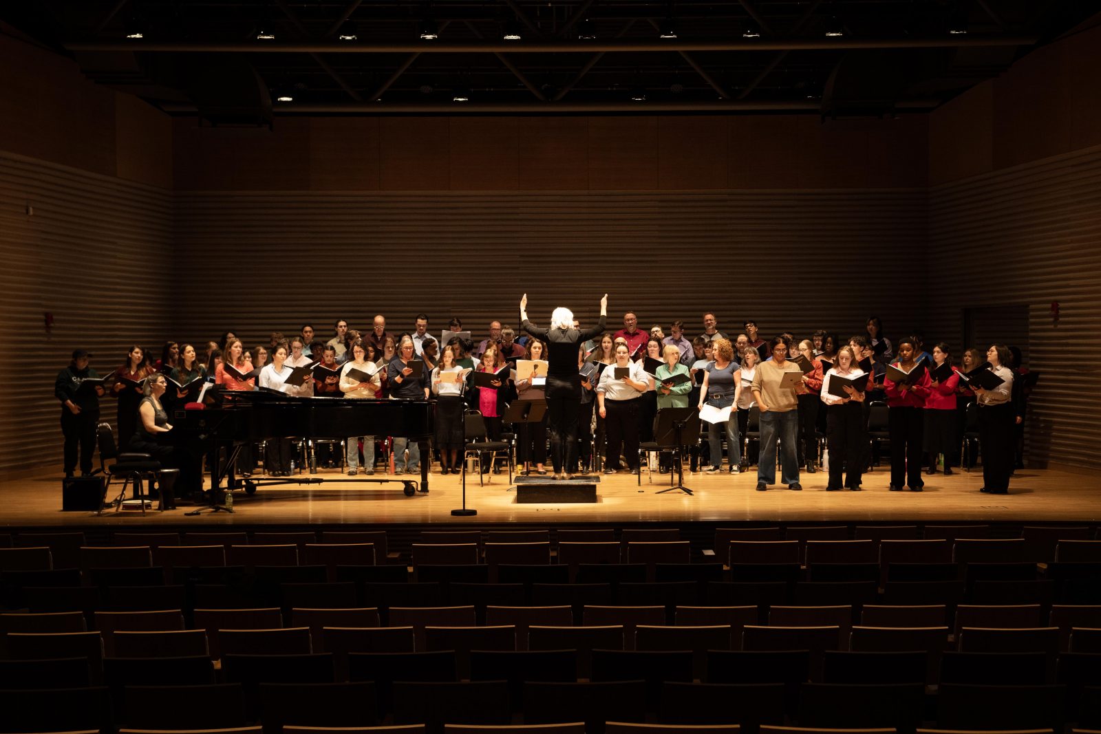 A conductor leads a large group of people who are singing on stage in a professional theatre with auditorium seating in the foreground.