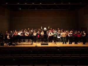 A conductor leads a large group of people who are singing on stage in a professional theatre with auditorium seating in the foreground.