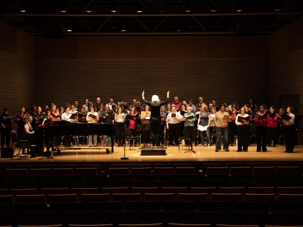 A conductor leads a large group of people who are singing on stage in a professional theatre with auditorium seating in the foreground.