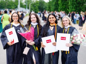 Four university graduates pose in gowns holding flowers and diplomas at a Convocation ceremony.