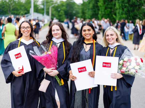 Four university graduates pose in gowns holding flowers and diplomas at a Convocation ceremony.