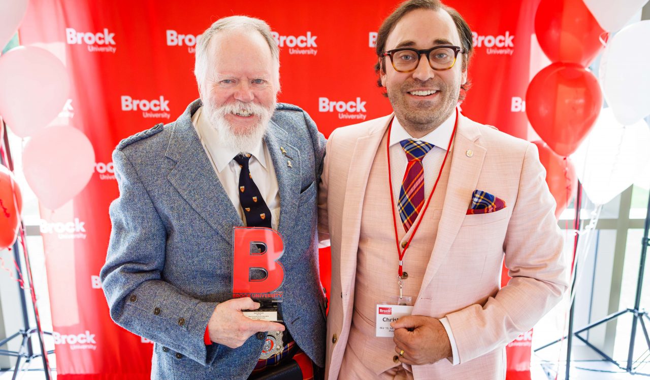 Two men in suits stand in front of a red backdrop.