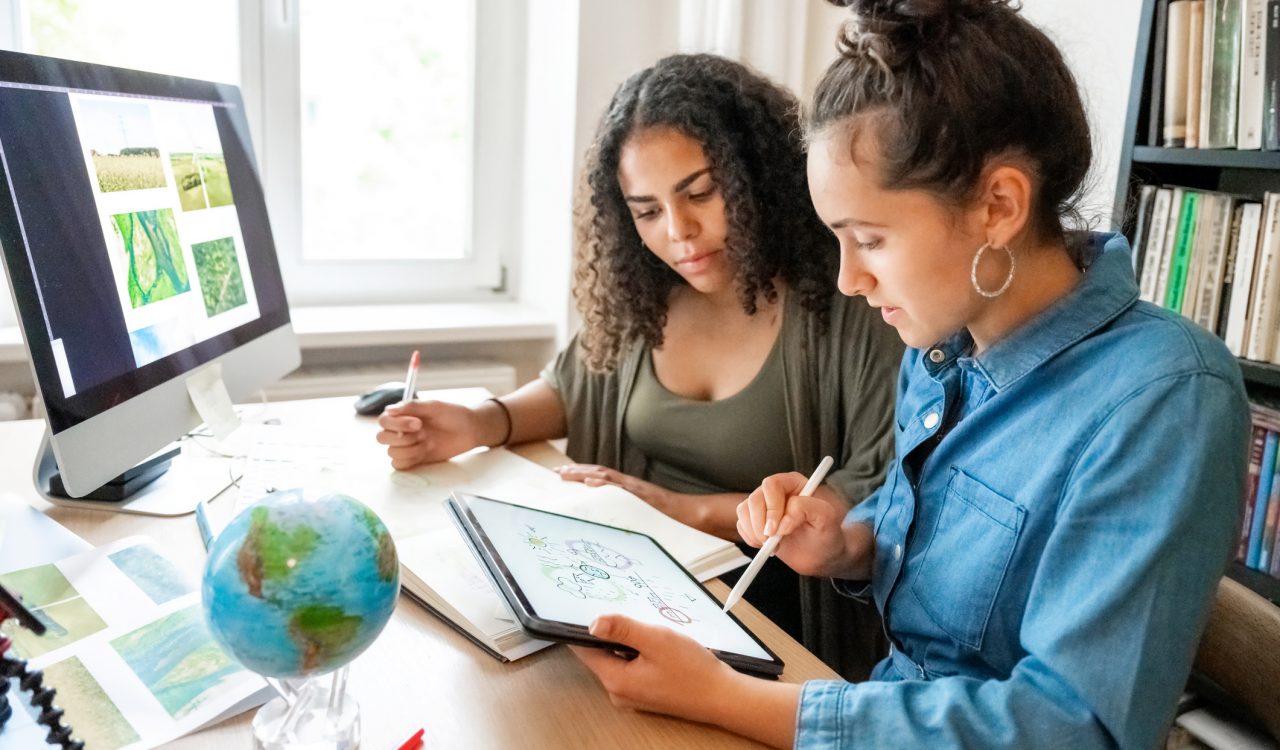 Two women look over documents and an iPad while sitting at a desk.