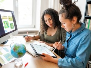 Two women look over documents and an iPad while sitting at a desk.