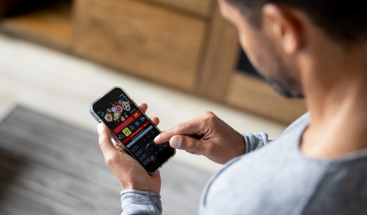 A man, standing indoors, holds a mobile phone in one hand and touches the screen with his index finger.