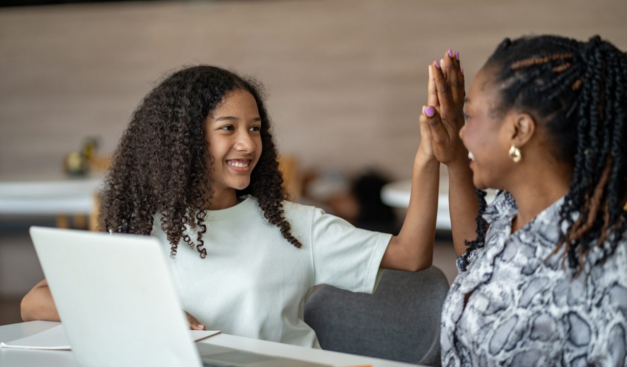 A smiling young person sits at a table with an open laptop, raising a hand to high-five an adult seated beside them. The pair appear engaged in a positive conversation in a bright indoor space.