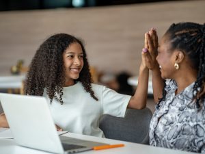 A smiling young person sits at a table with an open laptop, raising a hand to high-five an adult seated beside them. The pair appear engaged in a positive conversation in a bright indoor space.