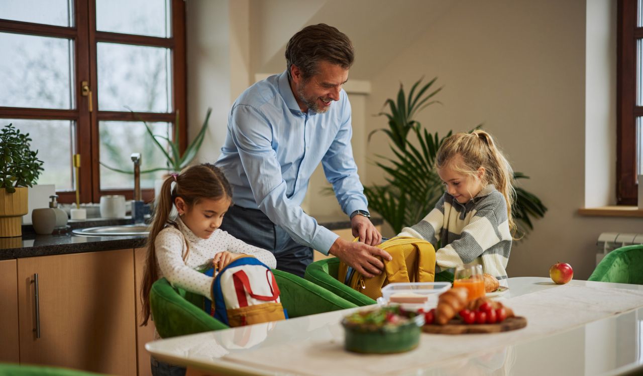 A father helps his two daughters pack their backpacks at the kitchen table.