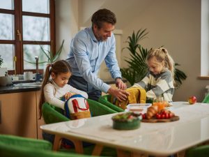 A father helps his two daughters pack their backpacks at the kitchen table.