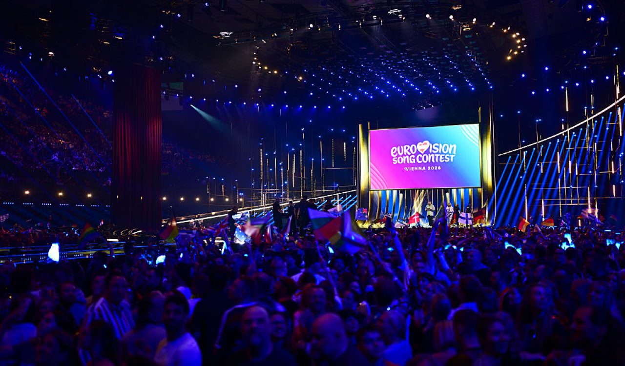 An auditorium packed with people looking on to a stage. The room is filled with concert lighting making it colourful throughout.