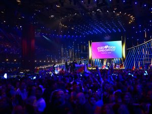 An auditorium packed with people looking on to a stage. The room is filled with concert lighting making it colourful throughout.