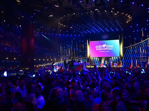 An auditorium packed with people looking on to a stage. The room is filled with concert lighting making it colourful throughout.