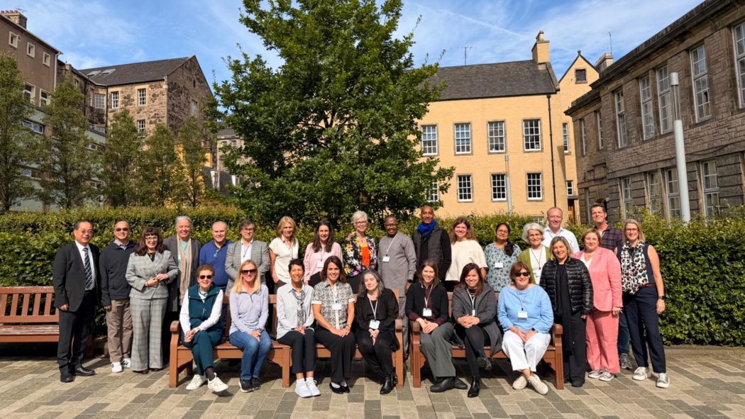 A large group of people pose for a photo, with some seated on benches, outside on a sunny day.