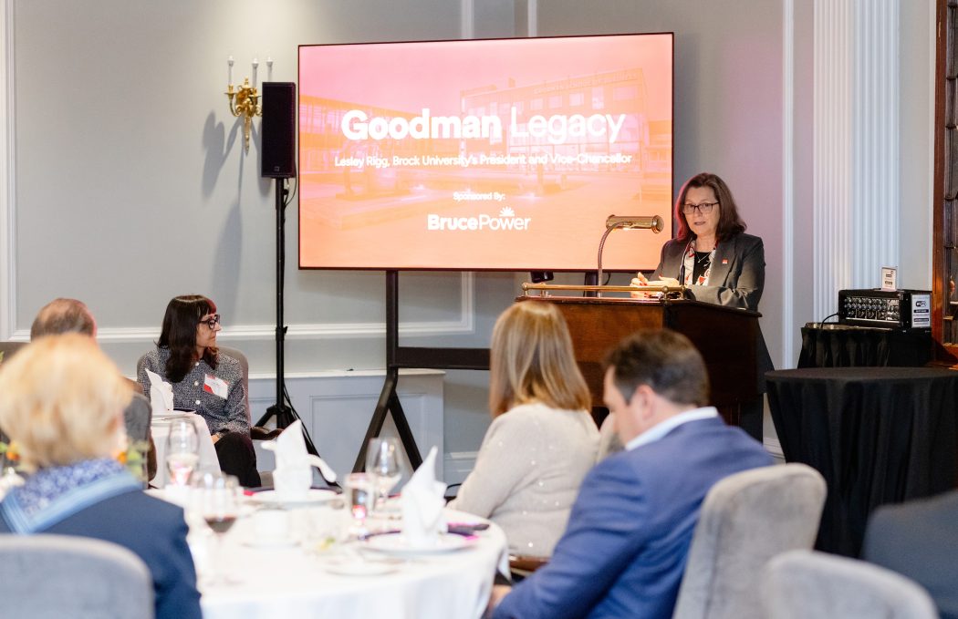 A women stands at the podium in front of a TV screen that reads Goodman Legacy as she speaks to tables full of guests. 