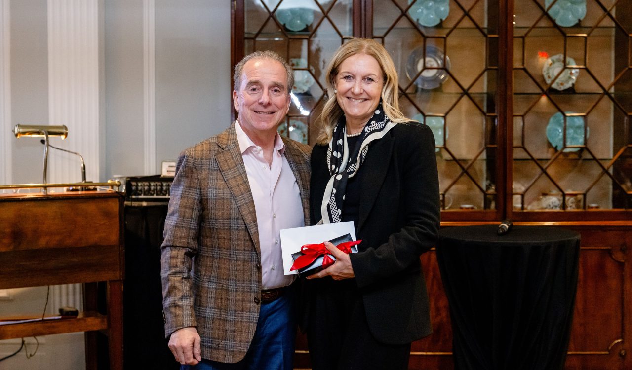A man in business attire passes a black gift box with a red bow to woman with business attire in front of a wall of cabinets with china.