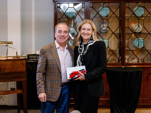 A man in business attire passes a black gift box with a red bow to woman with business attire in front of a wall of cabinets with china.