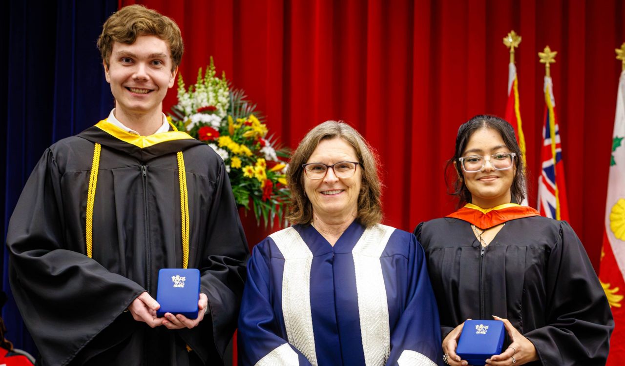 Three people in academic robes pose for a photo on stage during a university graduation ceremony. Two are holding medals.