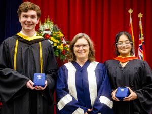 Three people in academic robes pose for a photo on stage during a university graduation ceremony. Two are holding medals.